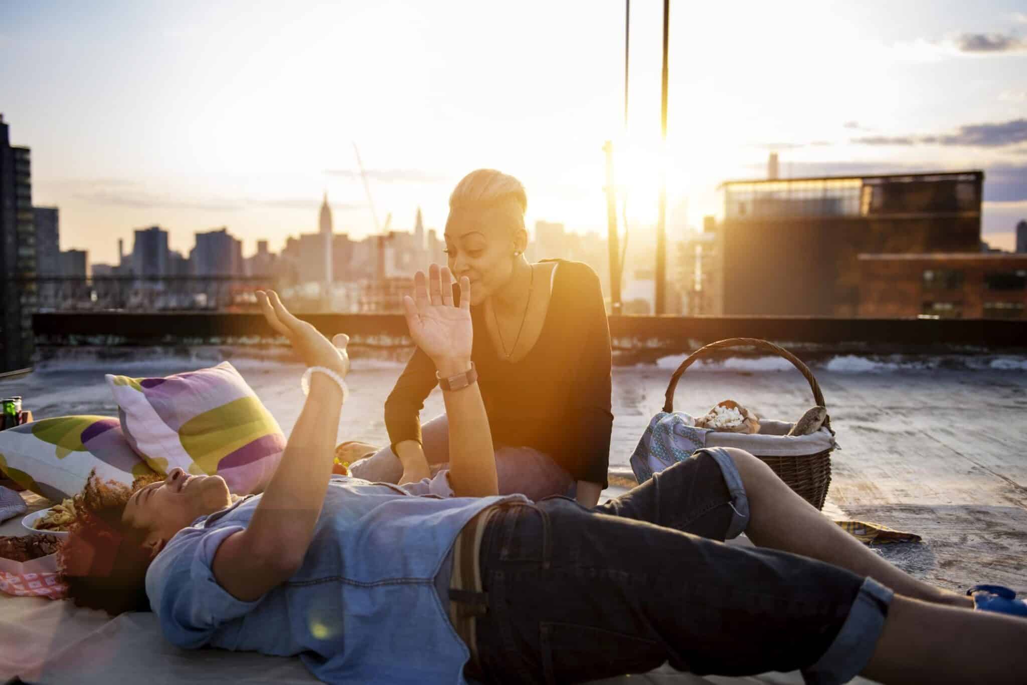 Man lying besides woman sitting on rooftop against sky during sunset