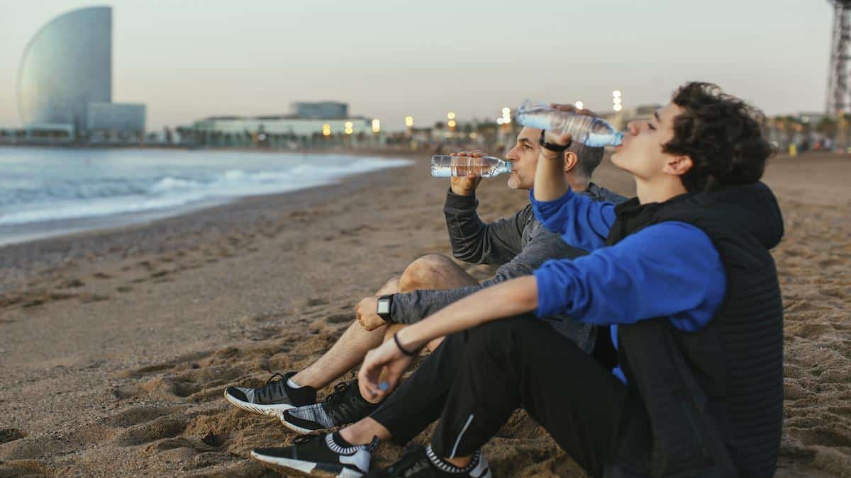 ​Two men drink bottled water on a beach in Barcelona, Spain.