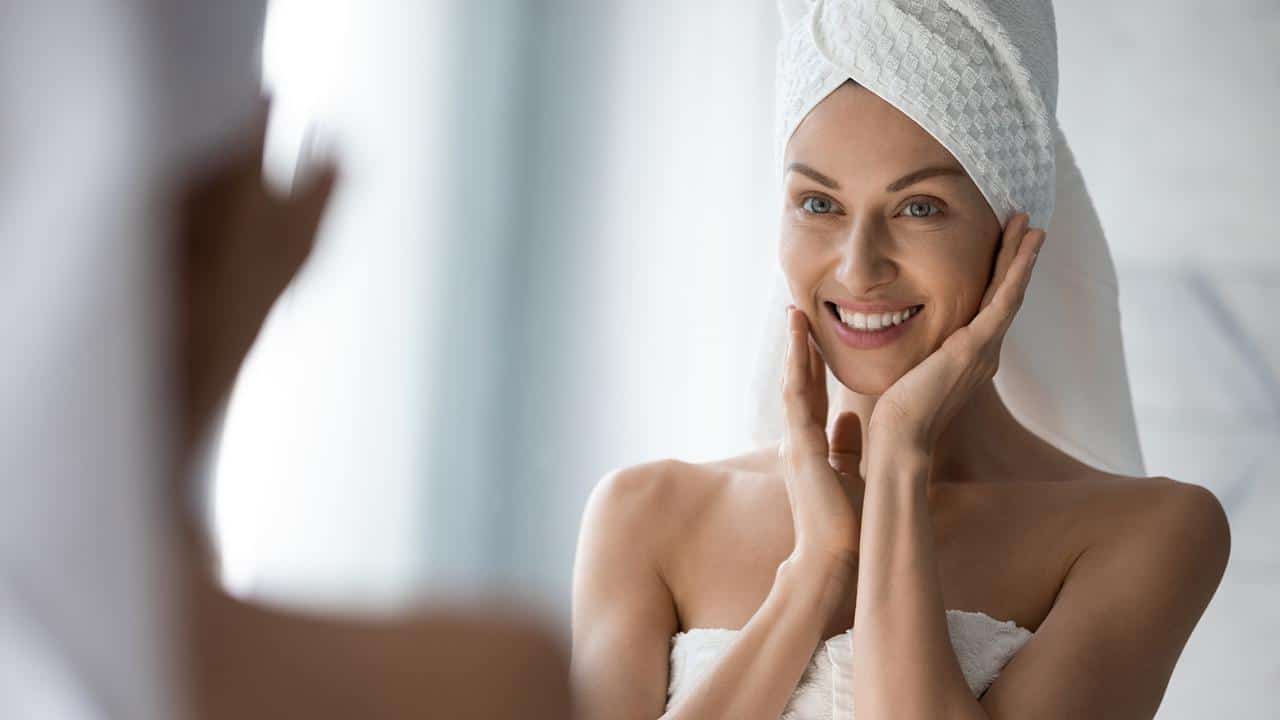 Woman looks in mirror touches moisturized healthy skin feels satisfied