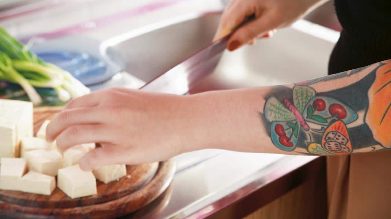 Woman chopping tofu in kitchen with ornate tattooed arm