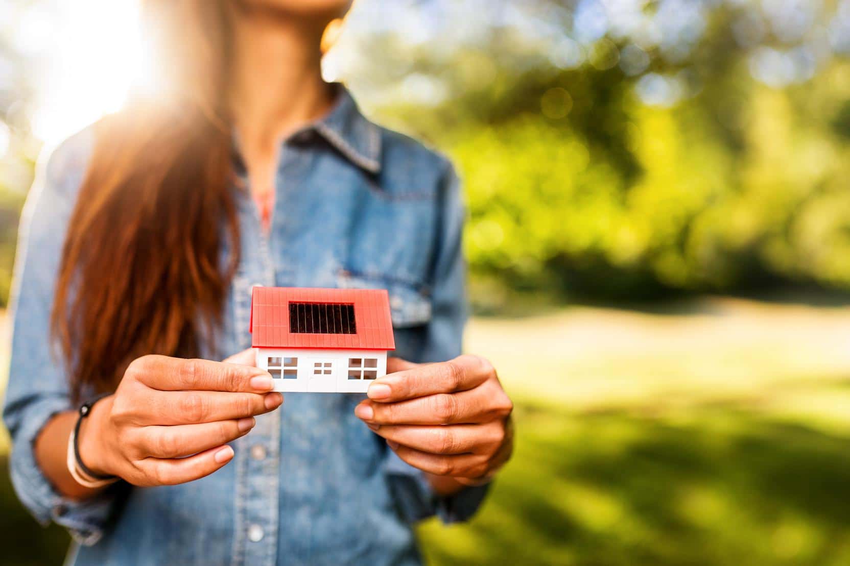 Woman presenting a model of a toy house with solar cells and a sunlit lawn behind her