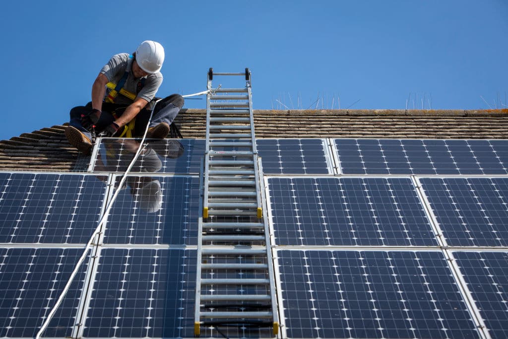 A maintenance person uses a ladder and harnesses to install equipment around a solar panel array on the roof of a house to stop birds nesting underneath