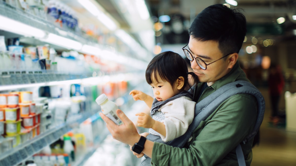 A man carrying his daughter in a supermarket reads a food label.