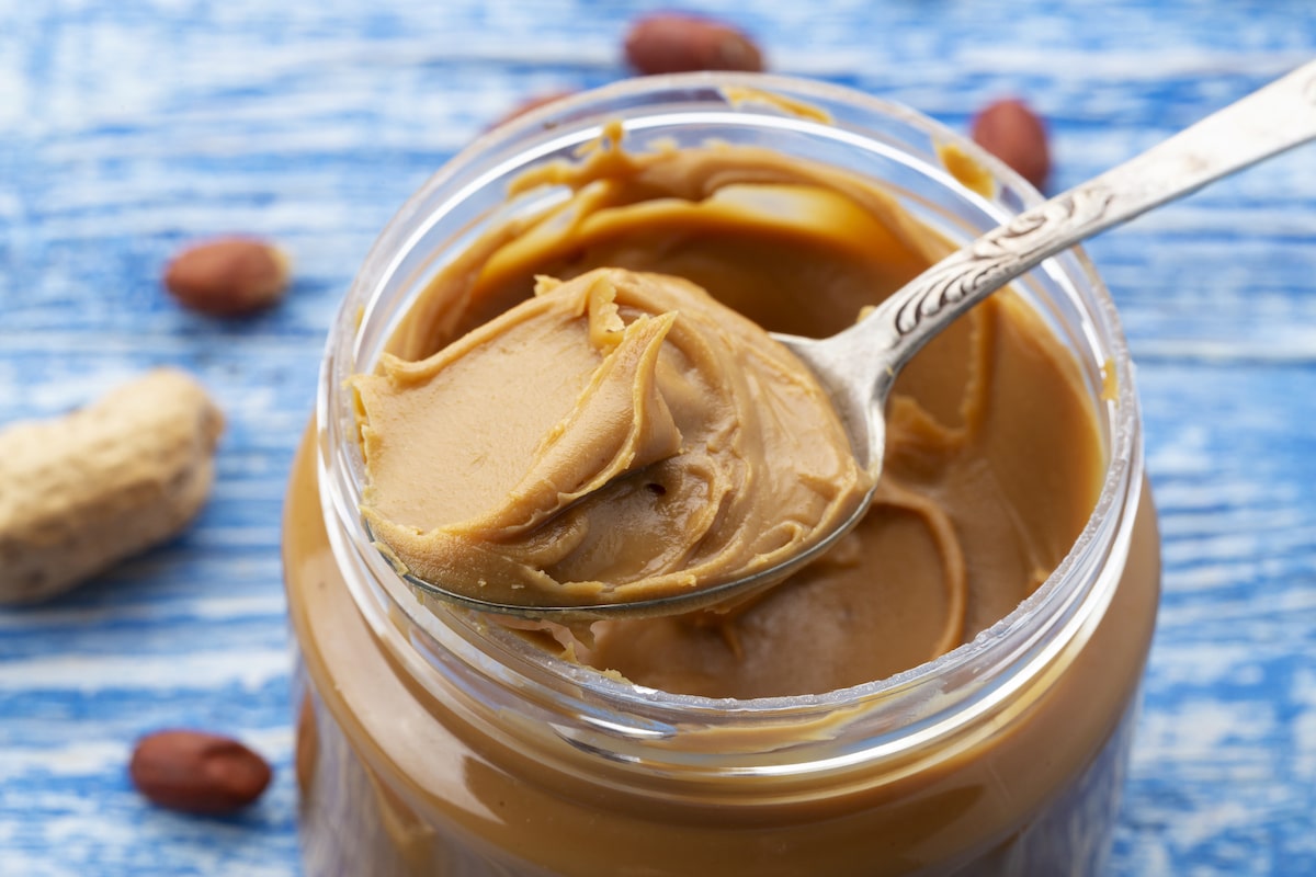 Peanut butter in an open jar and peanuts scattered on a blue table
