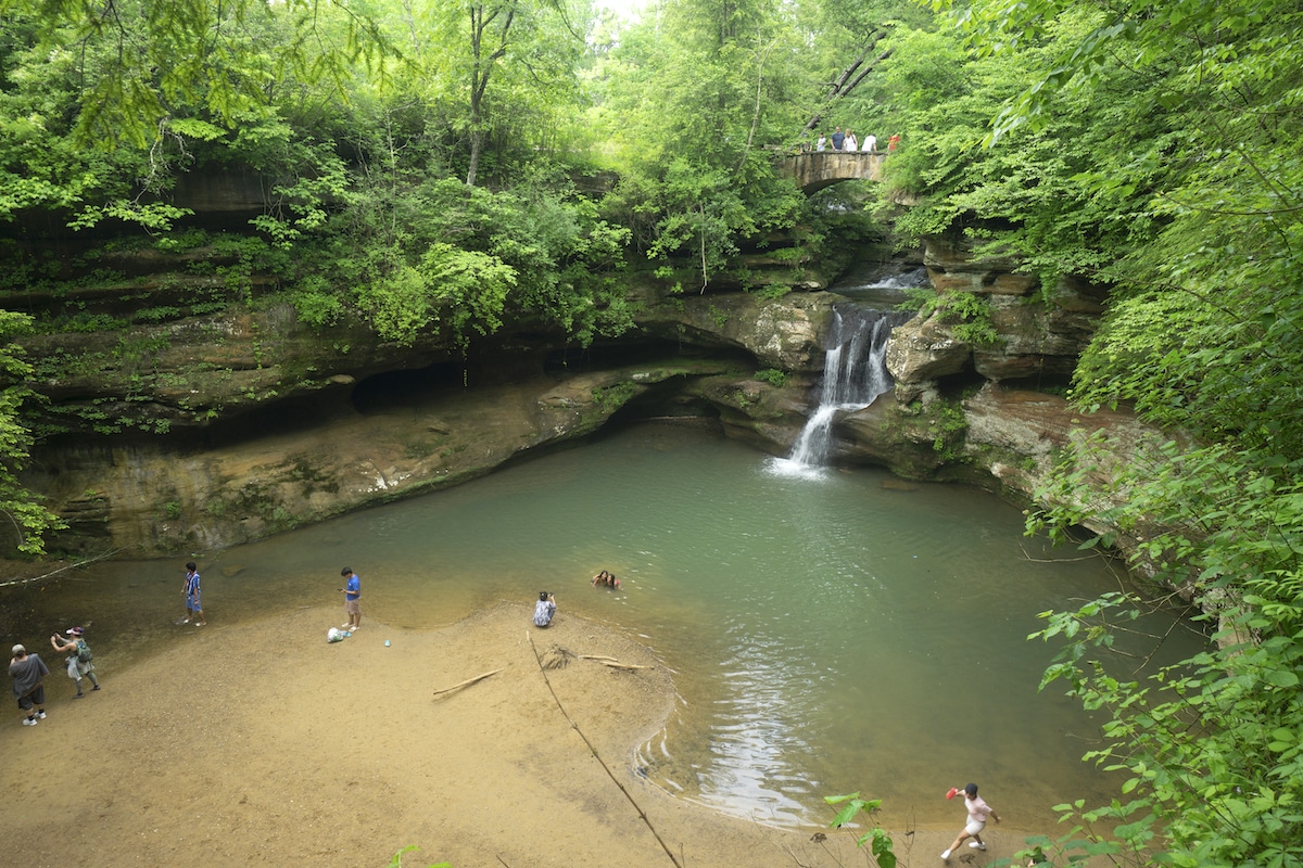 People enjoy a summer visit to Hocking Hills State Park in Ohio in an area with a waterfall, bridge, and pool.
