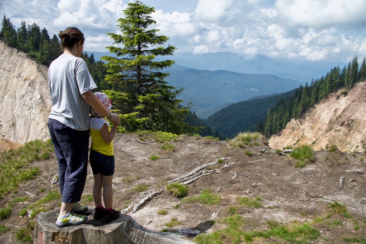 A mother and daugher standing on a tree stump look out over a mountain landscape in Romania
