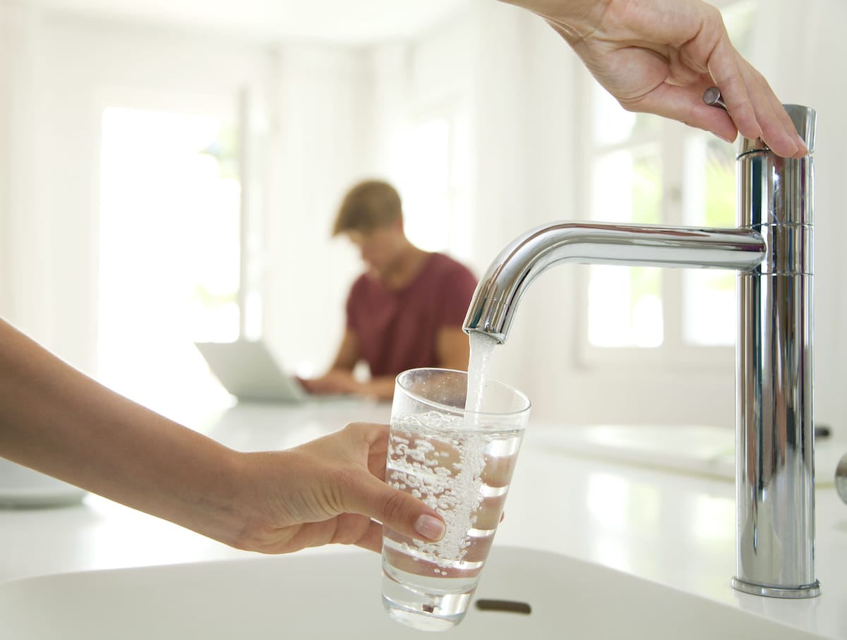 A person fills a drinking glass with water from a kitchen faucet while another person works at home in the background