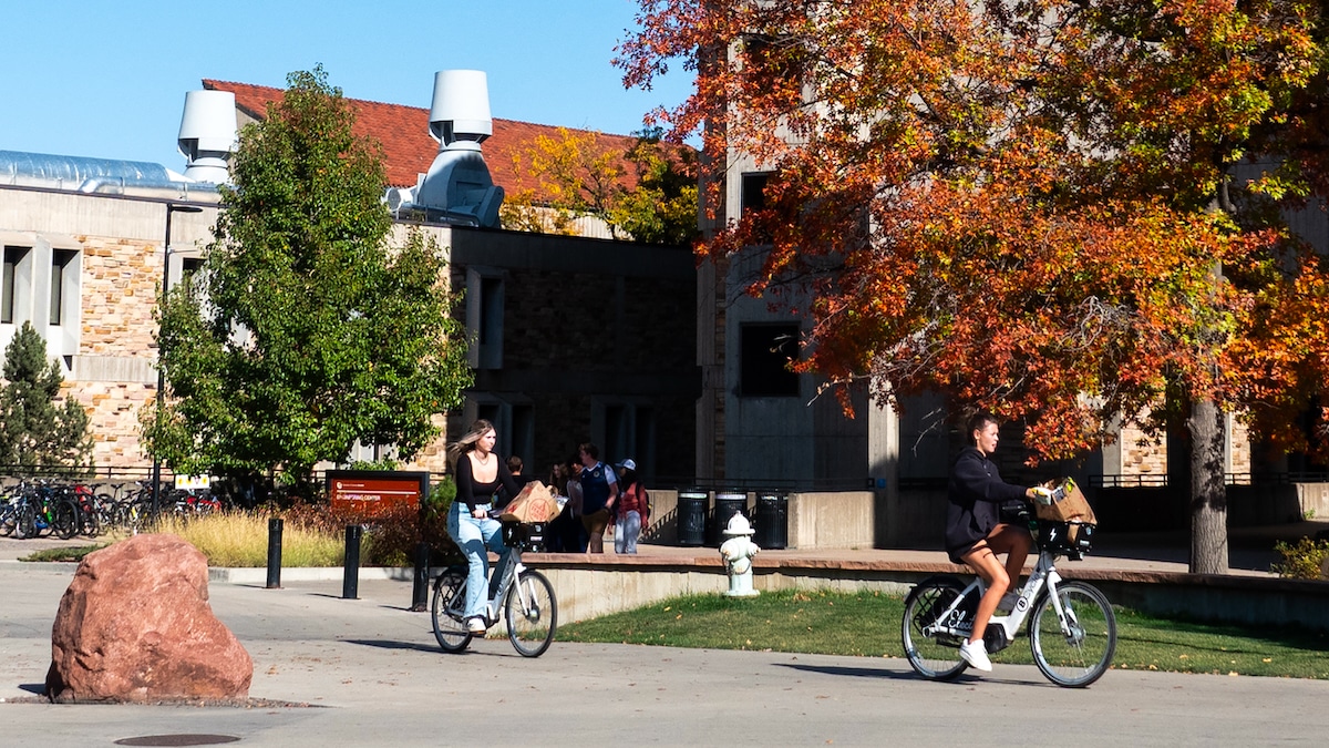 Two women bicyclists with groceries ride e-bikes on the University of Colorado, Boulder campus